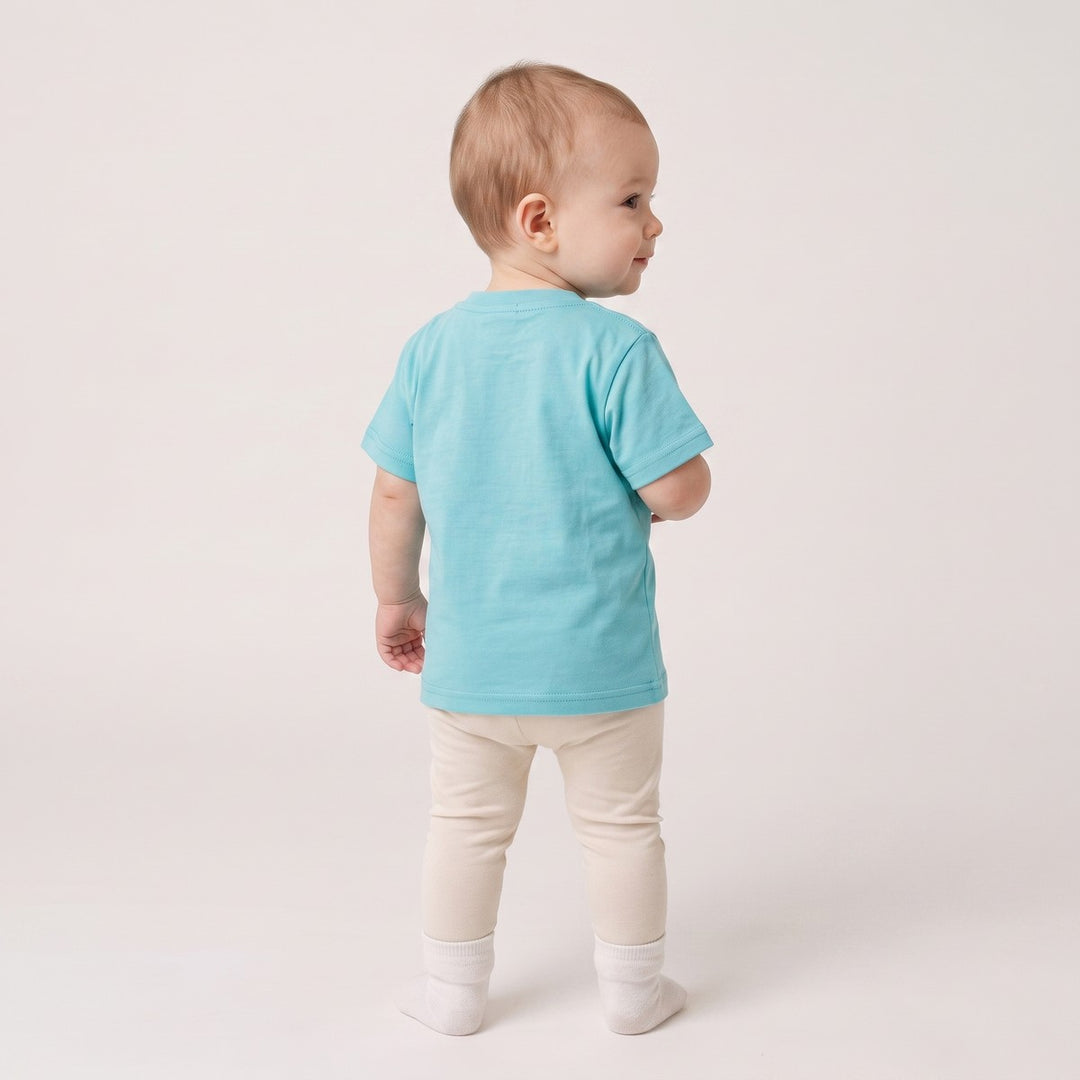 Back view of a baby wearing a turquoise short-sleeve t-shirt and light beige pants with white socks, standing on a white