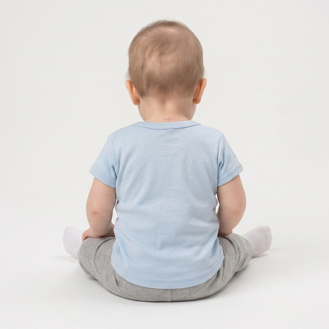 Back view of a baby wearing a light blue Elephant T-Shirt and gray pants, standing on a clean white studio background.